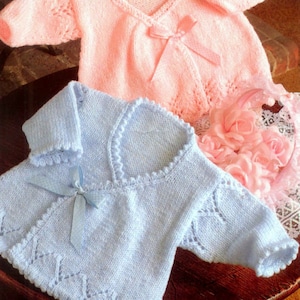 May include: Two knitted baby sweaters, one light pink and one light blue, with a decorative bow on each. The sweaters have a delicate lace pattern and are made with soft yarn.
