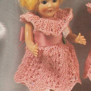 May include: A pink crocheted dress with a white lace trim and a pink satin sash. The dress is worn by a doll with blonde hair and blue eyes.