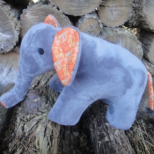 Op de afbeelding: Een grijs pluche olifantenspeelgoed met oranje stoffen oren. De oren zijn versierd met kleine olifanten. Het speelgoed zit op een stapel hout.