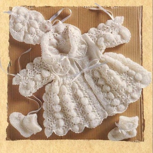 May include: A white knitted baby outfit with a jacket, hat, and booties. The outfit is made with a delicate stitch and has a lacy design.
