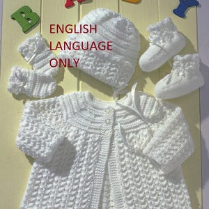 Puede incluir: Conjunto de bebé de punto blanco que incluye un gorro, una chaqueta y botines. El conjunto está colocado sobre un fondo de madera clara con las palabras "ENGLISH LANGUAGE ONLY" impresas en la imagen.