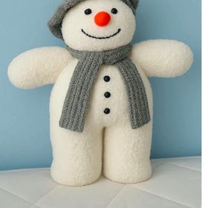 May include: A soft snowman toy, wearing a grey knitted hat and scarf. The snowman has a red nose, black button eyes, and a stitched smile. It stands on a white surface against a light blue background.