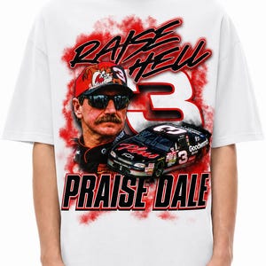 K&ouml;nnte beinhalten: Ein wei&szlig;es T-Shirt mit einem Grafikdruck eines Rennfahrers mit Sonnenbrille und Kappe, einem schwarzen Rennwagen mit der Nummer 3 und roten Raucheffekten. Der Text RAISE HELL und PRAISE DALE ist in fetten schwarzen und roten Buchstaben aufgedruckt.