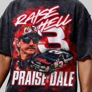 May include: A black and gray t-shirt featuring a graphic of a race car, a portrait of a man wearing sunglasses and a hat, and the text "RAISE HELL PRAISE DALE" in white and red. The number "3" is prominently displayed.