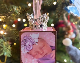 May include: A pink wooden cube ornament with a photo of a baby girl wearing a flower headband. The ornament has a silver crown on top and a pink ribbon for hanging.