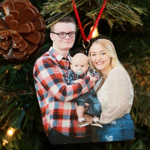 May include: A Christmas ornament featuring a family portrait. A man in a red and black plaid shirt holds a baby in a blue and white plaid shirt. A woman in a white sweater and blue jeans stands next to the man. The baby is wearing a blue pacifier.