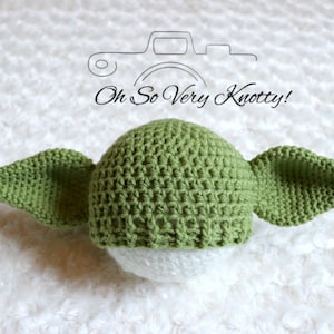 Puede incluir: Sombrero de bebé de ganchillo verde con orejas grandes, que se asemeja a un personaje ficticio popular. El sombrero está sobre una superficie blanca. "Oh So Very Knotty!" está escrito en negro sobre el sombrero.