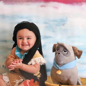 May include: A smiling baby wearing a crocheted Native American-inspired outfit with a black wig. A plush dog with a blue collar and a gold acorn charm sits nearby. The background features a watercolor sky.