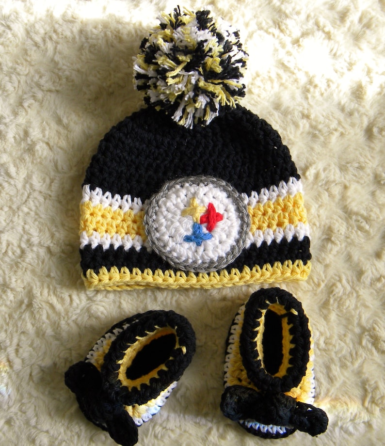May include: Black, yellow, and white crocheted baby hat with a pom-pom and matching booties. The hat features a Pittsburgh Steelers logo.