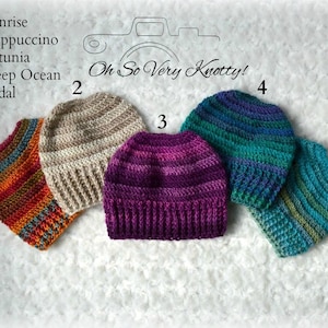 May include: Five crocheted hats in different colors and patterns, labeled with numbers 1 through 5. The hats are arranged in a semi-circle on a white fuzzy surface. The text "Oh So Very Knotty!" is written above the hats.