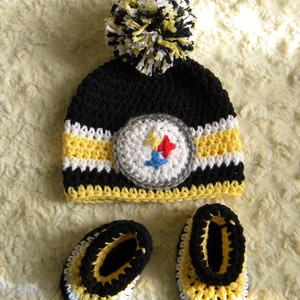 May include: A black, yellow, and white crocheted baby hat with a pom-pom on top and a Pittsburgh Steelers logo. The hat is paired with matching booties.