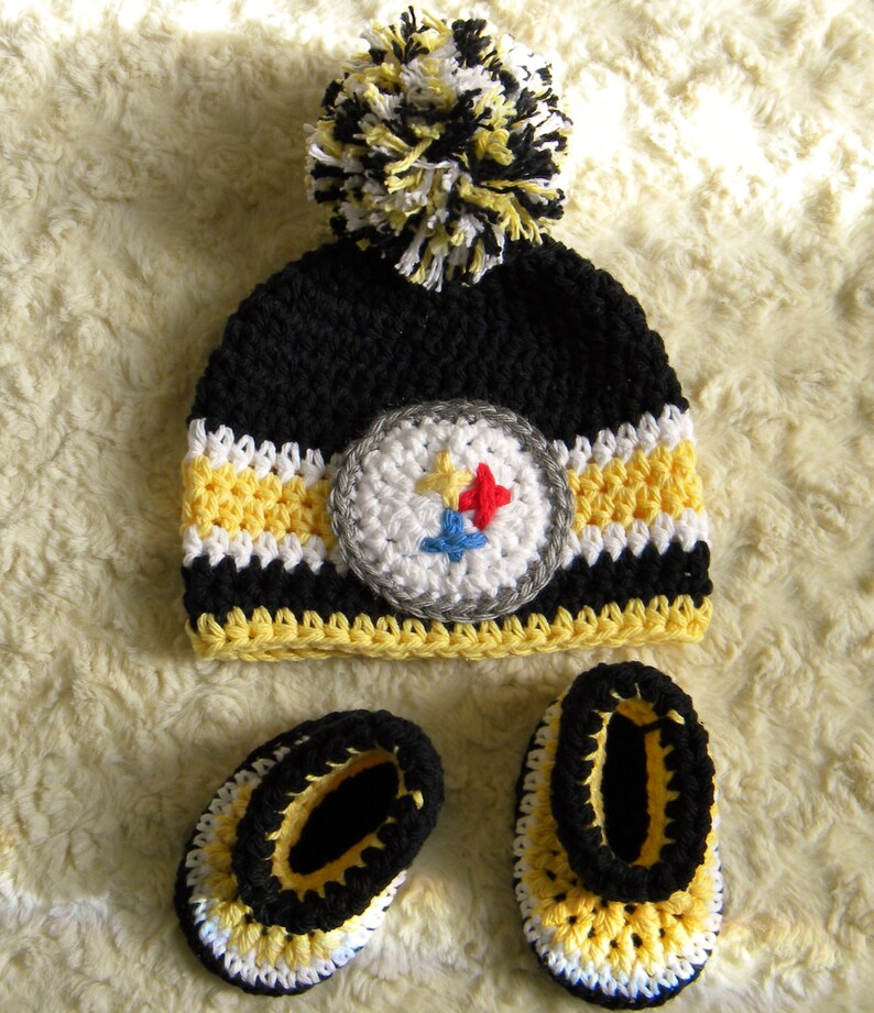 May include: A black, yellow, and white crocheted baby hat with a pom-pom on top and a Pittsburgh Steelers logo. The hat is paired with matching booties.