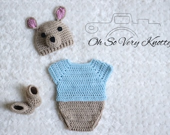 May include: A crocheted baby outfit in blue and brown with a matching crocheted kangaroo hat. The hat has pink ears and a brown nose. The outfit includes a bodysuit and booties. The text "Oh So Very Knotty!" is visible in the image.
