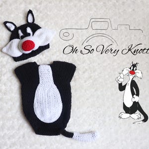 Puede incluir: Un conjunto de ganchillo en blanco y negro con temática de gato, que incluye un gorro con orejas y nariz roja, y un body con una mancha blanca en el vientre y una cola con la punta blanca. El texto Oh So Very Knotty! aparece junto a un dibujo.