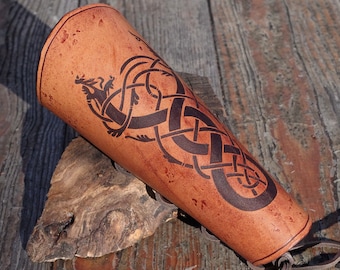 May include: A brown leather arm guard with a black Celtic knot design and a dragon motif. The arm guard has a strap for fastening.