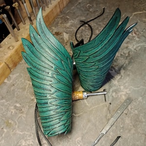 May include: Handmade teal leather wing arm cuffs with detailed feather texture. The cuffs are connected by a black cord and feature a wooden handle. A scalpel and tools are in the background.
