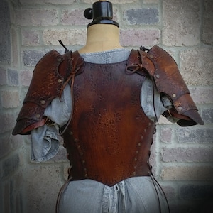 May include: Brown leather armor with a textured finish. The armor includes a chest piece and shoulder pads. The armor is displayed on a mannequin against a brick wall.