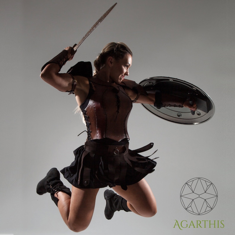 May include: A woman wearing a brown leather bodice and a black skirt jumps while holding a sword and a round shield. The shield is silver and has a raised rim. The woman is wearing black boots and has her hair pulled back.