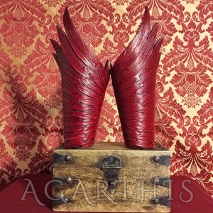 Könnte beinhalten: Ein Paar rote Lederflügel mit einem detaillierten, strukturierten Design. Die Flügel werden auf einer Holzkiste mit einem Metallverschluss präsentiert.