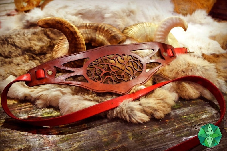May include: A brown leather belt with a decorative cutout design. The belt has a red strap and a metal buckle. The belt is lying on a fur-covered surface.