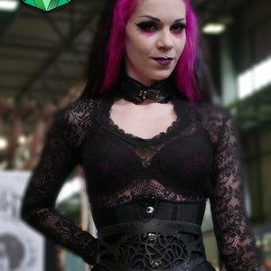 May include: A woman with pink hair and dark makeup is wearing a black lace top, a black corset with a decorative design, and a black choker with a silver buckle. She is also wearing a pair of black sunglasses.