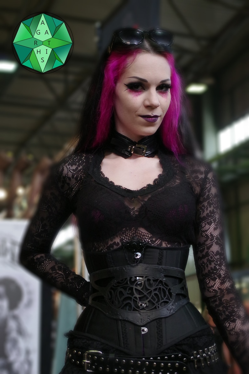 May include: A woman with pink hair and dark makeup is wearing a black lace top, a black corset with a decorative design, and a black choker with a silver buckle. She is also wearing a pair of black sunglasses.