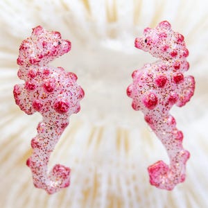 May include: Two pink seahorse-shaped earrings with red accents and glitter. The earrings are set against a white, textured background. The seahorses have a whimsical, decorative design.