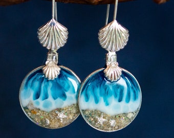 May include: A pair of silver dangle earrings with a blue and white seashell design. The earrings feature a small seashell charm and a larger round charm with a blue and white seascape design. The seascape design includes a white sandy beach with gold glitter and a blue ocean with white waves.