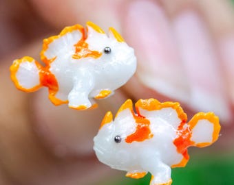 May include: A pair of small, white fish-shaped earrings with orange and white accents. Each earring has a small, silver-colored stud. The earrings are held in a person's hand against a blurred green background.