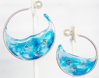 May include: A pair of silver hoop earrings with blue and white resin inserts depicting a wave with small silver fish swimming inside.