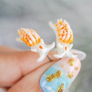 Puede incluir: Un par de pendientes de babosa de mar blancos y naranjas. Los pendientes están sostenidos por una mano con un diseño de uñas pintadas con adornos dorados de caballito de mar, conchas marinas y estrellas de mar.