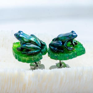 May include: A pair of stud earrings featuring small, iridescent blue-green frogs perched on top of textured, glittery green lily pads. The earrings have silver-toned metal posts and backings, showcasing a detailed, three-dimensional design.
