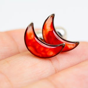 May include: A pair of crescent moon earrings with a red and orange marbled design. The earrings are made of metal and have a silver finish.