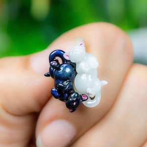 May include: A small ring with two animal figures, one dark blue and the other white. The figures are intertwined. The ring is held in a hand against a blurred green background. The ring's design is highly detailed.