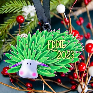 Puede incluir: Adorno navide&ntilde;o decorativo con un dise&ntilde;o de oveja estilizada, follaje verde y detalles morados. El adorno tiene el nombre "EDDIE" y el a&ntilde;o "2023" en letras doradas. Cuelga de una cinta negra.