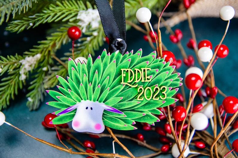Puede incluir: Adorno navide&ntilde;o decorativo con un dise&ntilde;o de oveja estilizada, follaje verde y detalles morados. El adorno tiene el nombre "EDDIE" y el a&ntilde;o "2023" en letras doradas. Cuelga de una cinta negra.