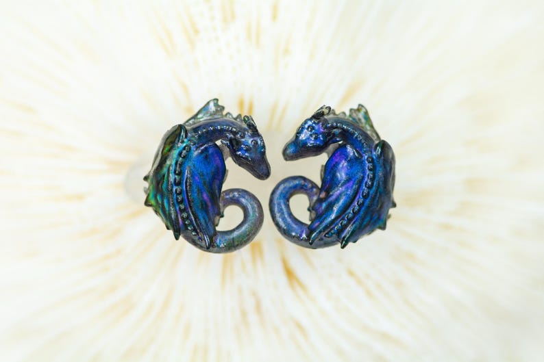 May include: Two iridescent blue dragon earrings. The dragons are curled up in a sleeping position. The earrings are made of a shiny, metallic material.