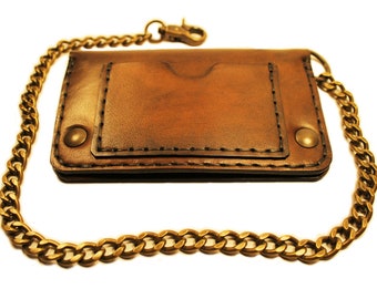 Pode incluir: Uma carteira de couro marrom com uma corrente presa. A carteira tem uma aba com um bolso e é costurada com fio marrom. A corrente é de latão e tem um fecho.