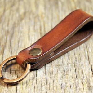 May include: A brown leather key fob with a brass-colored split ring and a brass rivet. The key holder is made of a folded piece of leather. The background is a wooden surface.