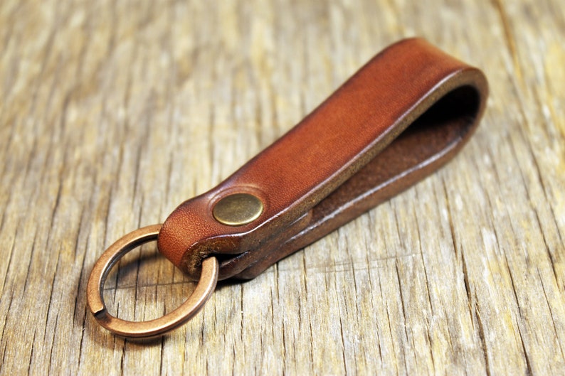 May include: A brown leather key fob with a brass-colored split ring and a brass rivet. The key holder is made of a folded piece of leather. The background is a wooden surface.