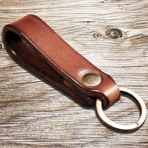 May include: A brown leather keychain with a brass rivet and a silver-colored key ring. The leather is a warm, medium brown color. The keychain is lying on a wooden surface.