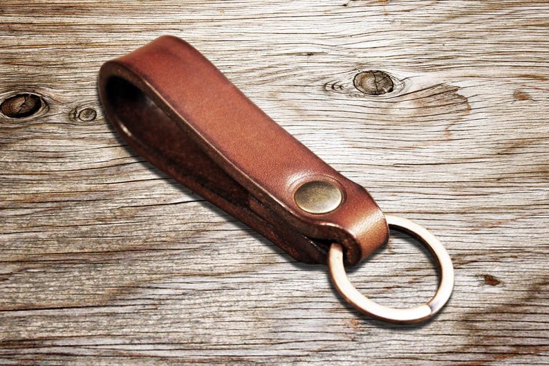 May include: A brown leather keychain with a brass rivet and a silver-colored key ring. The leather is a warm, medium brown color. The keychain is lying on a wooden surface.