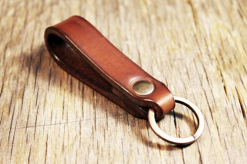 May include: A brown leather keychain with a brass-colored rivet and a metal ring. The keychain is made of a single piece of folded leather. The leather has a rich, warm color and a smooth texture.