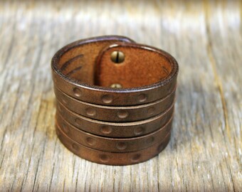 May include: A brown leather cuff bracelet with a hammered texture and a brass rivet. The bracelet is made of multiple layers of leather and is designed to be worn on the wrist.