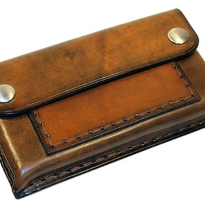 Peut inclure: Un portefeuille en cuir marron avec une fermeture à rabat et deux boutons-pression argentés. Le portefeuille a une petite poche sur le devant avec un bord cousu.