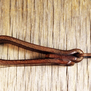 May include: A brown leather key fob with a copper-colored key ring. The key fob is looped and knotted, with a rich, textured appearance. The background is a light brown, weathered wood.