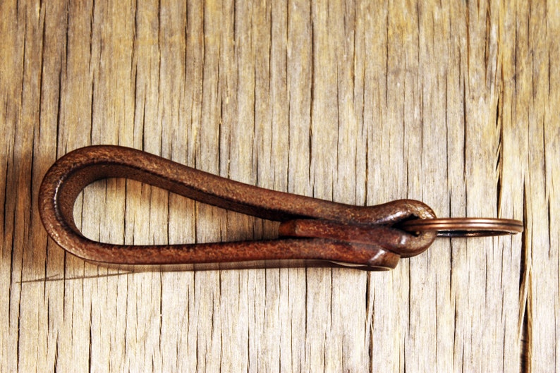 May include: A brown leather key fob with a copper-colored key ring. The key fob is looped and knotted, with a rich, textured appearance. The background is a light brown, weathered wood.