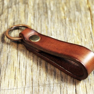 May include: A brown leather key fob with a brass rivet and a bronze key ring. The key holder is made of a folded strip of leather. The key fob is on a wooden surface.