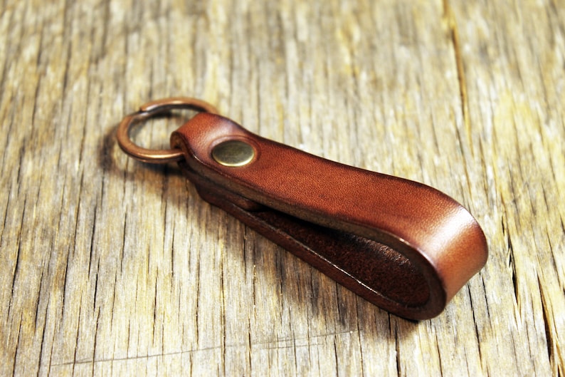 May include: A brown leather key fob with a brass rivet and a bronze key ring. The key holder is made of a folded strip of leather. The key fob is on a wooden surface.
