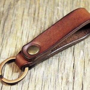 May include: A brown leather keychain with a brass-colored key ring and a brass button closure. The leather is a rich, warm brown color, and the key ring is attached to a loop of the same leather. The keychain is shown on a wooden surface.